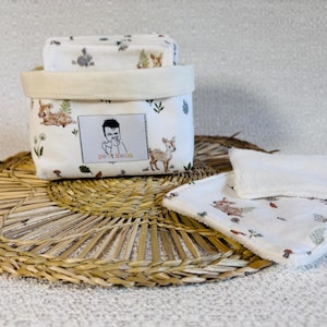 Pode incluir: Uma cesta de tecido branco e panos a condizer com uma estampa de animais da floresta. A cesta tem uma borda creme e uma etiqueta com um desenho animado e o texto "petit deco". Os itens estão em um tapete tecido.