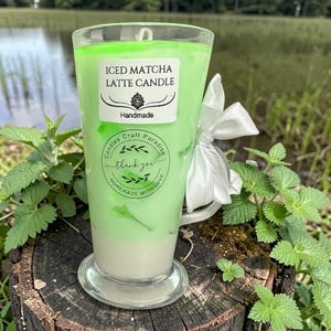 May include: A handmade Iced Matcha Latte candle in a clear glass with a handle. The candle has layers of white and green wax, and is decorated with a white ribbon. The label reads "Iced Matcha Latte Candle".
