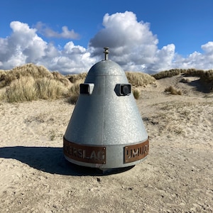 Op de afbeelding: Een metaalgrijze, kegelvormige structuur met twee rechthoekige openingen en een kleine antenne bovenop, staand op een zandstrand. De structuur heeft tekst op een band rond de basis. De achtergrond toont zandduinen, gras en een blauwe lucht met wolken.