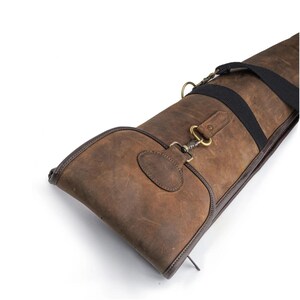 Puede incluir: Un estuche para armas de cuero marrón con ribete marrón oscuro y una correa negra. El estuche tiene un cierre de solapa con un cierre color latón y un parche de cuero. Diseñado para transportar un arma larga.