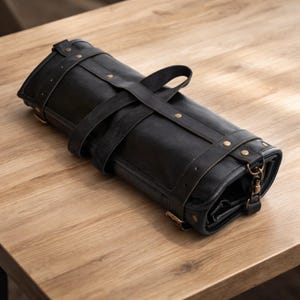 Puede incluir: Un estuche enrollado de cuero negro para cuchillos, con remaches y hebillas de color latón. Tiene un asa y correas para asegurar el contenido. Diseñado para guardar y proteger herramientas culinarias.