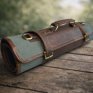 Puede incluir: Un estuche enrollado para cuchillos de cuero marrón y lona verde oliva. Tiene un asa de cuero y herrajes de color latón. El estuche se asegura con correas y hebillas de cuero. El fondo es una superficie de madera borrosa.