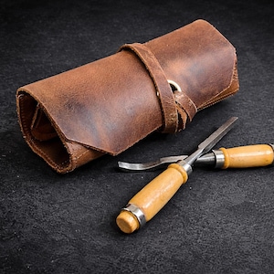 May include: A rolled brown leather tool roll with a strap and two wood-handled chisels. The chisels have metal blades and are lying on a dark surface. The leather roll is closed with a strap and metal ring.