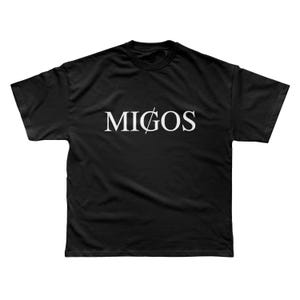 Op de afbeelding: Zwart T-shirt met het woord "MIGOS" in witte letters, met een streep door de "O". Het shirt heeft een ronde hals en korte mouwen. De tekst staat in het midden van de voorkant van het shirt.