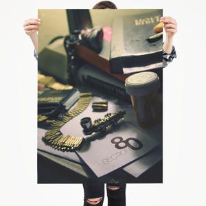 May include: A person holding a poster with a still life of gold jewelry, a pill bottle, books, and a small pipe. The poster also features the number 80 and the word "SECTION".