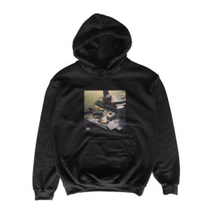 Peut inclure: Sweat à capuche noir avec une poche avant et une capuche à cordon. Le sweat-shirt présente un graphique rectangulaire d'un bureau avec divers objets, dont un sac, des papiers et des lunettes. Le graphique est dans des tons sourds.
