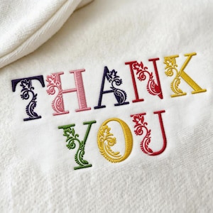 May include: White towel with the embroidered words "THANK YOU" in a variety of colors. The letters are decorated with floral designs. The towel is folded over at the top.