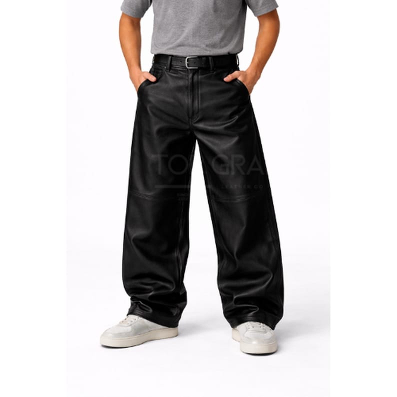 May include: Black leather pants with a wide leg design. The pants have a belt with a silver buckle and are worn with a gray t-shirt and white sneakers. The text "TO GRA LEATHER CO" is visible on the pants.
