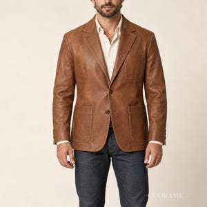 Op de afbeelding: Een lichtbruine leren blazer met een ingekeepte revers, twee opgestikte voorzakken, een borstzak en twee knopen aan de voorzijde. De blazer wordt gedragen over een lichtgekleurd overhemd en een donkere spijkerbroek van denim.