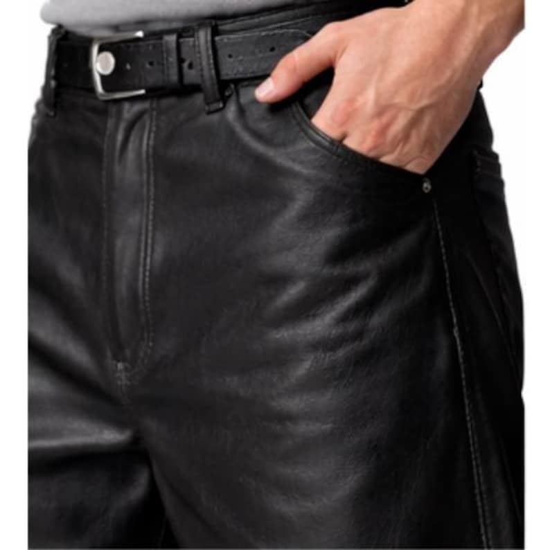 May include: Close-up of black leather pants with a matching belt. The pants have a classic five-pocket design and a straight leg cut. The belt is secured with a silver buckle. The person's hand is in the front pocket.
