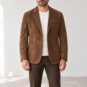 Puede incluir: Una americana de ante marrón con solapa de muesca, dos botones frontales y dos bolsillos de parche. La prenda se lleva sobre una camiseta blanca de cuello redondo, combinada con pantalones de color marrón oscuro. Texto CUORAME abajo a la derecha.