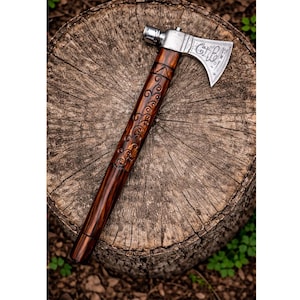 Puede incluir: Un hacha tomahawk artesanal con un mango de madera oscura y una cabeza plateada. El mango presenta intrincados diseños tallados. El hacha descansa sobre un tocón de árbol, mostrando su atractivo rústico.