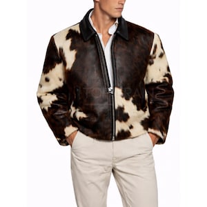 Può includere: Giacca in pelle di vacchetta marrone e bianca con colletto nero e cerniera frontale. La giacca ha un design classico ed è realizzata in un materiale morbido e strutturato. Il modello indossa una camicia bianca e pantaloni beige.