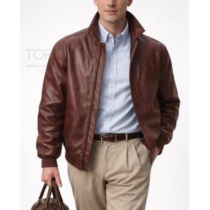 Può includere: Giacca in pelle marrone con zip e collo alto. La giacca ha polsini a coste e una cintura in pelle marrone. La persona indossa una camicia azzurra abbottonata e pantaloni beige. La persona tiene in mano una borsa in pelle marrone.