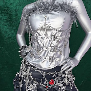 May include: Silver corset with chain and cross details, paired with a black skirt. The corset features buckles and a feathered trim. A spiked bracelet and a choker with a red cross complete the look. The overall style is gothic.