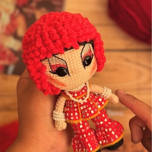Puede incluir: Muñeca de ganchillo hecha a mano con una peluca rizada roja, ojos negros grandes y un atuendo rojo con detalles cuadrados plateados. La muñeca tiene un collar y pulseras de perlas. La muñeca está siendo sostenida por una persona.