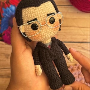 Puede incluir: Muñeca de ganchillo hecha a mano de una persona con traje negro, gafas y corbata rosa. La muñeca tiene pelo negro y tez clara. La muñeca está sujeta en una mano.