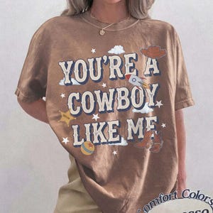 以下が含まれることがあります： 「YOU'RE A COWBOY LIKE ME」という文字がかすれたフォントでプリントされた茶色の半袖Tシャツ。グラフィックには、ロケット、カウボーイハット、星型のバッジ、ボール、カウボーイブーツのイラストが描かれており、小さな白い星が散りばめられています。