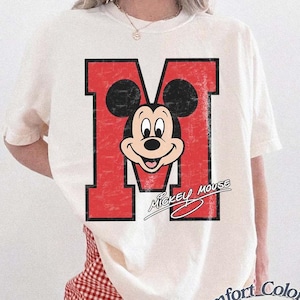 May include: Ivory-colored t-shirt featuring a distressed red letter "M" with a Mickey Mouse graphic. The shirt has the words "Mickey Mouse" in a script font below the graphic. The shirt is part of the Comfort Colors collection.