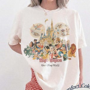 May include: Ivory-colored t-shirt featuring a colorful graphic of Disney characters and the Magic Kingdom castle. The text "Magic Kingdom Walt Disney World" is printed below the characters. The shirt is a Comfort Colors brand.