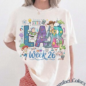 Op de afbeelding: Ivoor kleurig T-shirt met een kleurrijke "LAB Week 26" afbeelding met cartoonfiguren en wetenschappelijke illustraties. Het ontwerp bevat een microscoop, bekers en de woorden "Comfort Colors Ivory".