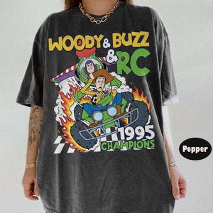 May include: Dark gray t-shirt featuring a graphic of Woody and Buzz Lightyear from Toy Story on a green RC car with flames. The text "Woody & Buzz & RC" and "1995 Champions" are also displayed.