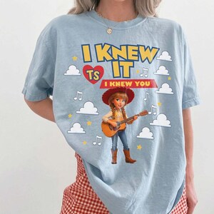 May include: A light blue t-shirt featuring a graphic of a cowgirl playing an acoustic guitar. The text reads I KNEW IT and I KNEW YOU, accompanied by a red heart, yellow stars, white clouds, and musical notes printed on the front of the garment.