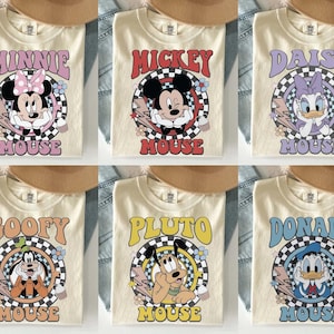 May include: Cream-colored t-shirts featuring retro designs of Disney characters: Minnie, Mickey, Daisy, Goofy, Pluto, and Donald. Each shirt has a character's name and image within a checkered circle, with lightning bolts and floral accents.