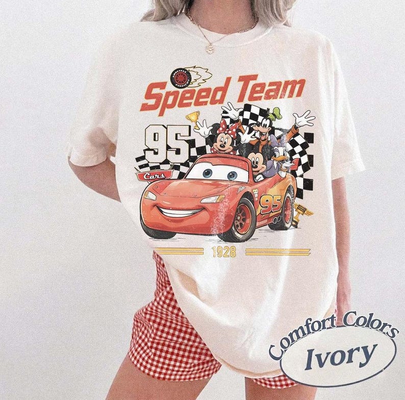 May include: Ivory-colored t-shirt featuring a graphic of Lightning McQueen from the Cars movie, along with Mickey and Minnie Mouse, Goofy, and Donald Duck. The shirt has the text "Speed Team 95 Cars" and "1928". The shirt is a Comfort Colors brand.