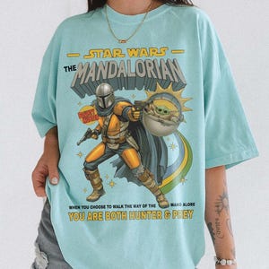 May include: A light blue t-shirt featuring a graphic print of The Mandalorian and Grogu. The text reads STAR WARS THE MANDALORIAN, BOUNTY HUNTER, and WHEN YOU CHOOSE TO WALK THE WAY OF THE MANDALORE YOU ARE BOTH HUNTER & PREY in yellow and black.