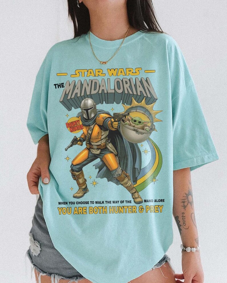 May include: A light blue t-shirt featuring a graphic print of The Mandalorian and Grogu. The text reads STAR WARS THE MANDALORIAN, BOUNTY HUNTER, and WHEN YOU CHOOSE TO WALK THE WAY OF THE MANDALORE YOU ARE BOTH HUNTER & PREY in yellow and black.