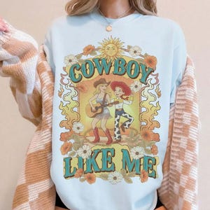 May include: A light blue crewneck sweatshirt featuring a graphic print of two cartoon characters, one playing a guitar, surrounded by a sun, flowers, and flames. The text COWBOY LIKE ME is printed in a teal western-style font above and below the characters.