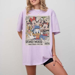 May include: A light purple t-shirt featuring a vintage-style graphic of Disney characters, including Mickey Mouse, Minnie Mouse, Donald Duck, and Goofy. The graphic reads "Disney World" and "2026" with the phrase "Where Dreams Come True".