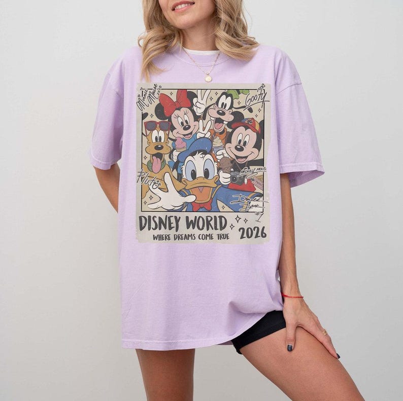 May include: A light purple t-shirt featuring a vintage-style graphic of Disney characters, including Mickey Mouse, Minnie Mouse, Donald Duck, and Goofy. The graphic reads "Disney World" and "2026" with the phrase "Where Dreams Come True".