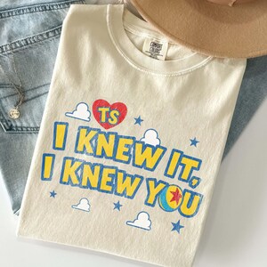 Puede incluir: Una camiseta de manga corta color crema con un coraz&oacute;n rojo que lleva las letras TS, texto azul que dice I KNEW IT, I KNEW YOU, y un gr&aacute;fico de una pelota con una estrella roja. El dise&ntilde;o incluye peque&ntilde;as estrellas azules y contornos de nubes blancas.