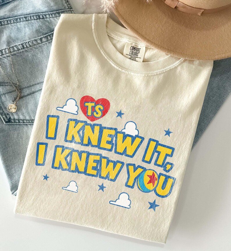 Puede incluir: Una camiseta de manga corta color crema con un coraz&oacute;n rojo que lleva las letras TS, texto azul que dice I KNEW IT, I KNEW YOU, y un gr&aacute;fico de una pelota con una estrella roja. El dise&ntilde;o incluye peque&ntilde;as estrellas azules y contornos de nubes blancas.