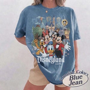 Pu&ograve; includere: Una t-shirt color blu jeans con una stampa grafica di Topolino, Minnie, Paperino, Pippo e Pluto davanti a un castello. Il testo Disneyland Est. 1955 &egrave; stampato sotto i personaggi. La maglietta presenta un girocollo.