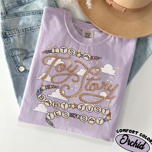 May include: A light purple t-shirt featuring a graphic design with the text "ITS A Toy Story BABY JUST YES SAY" written in rope-style lettering and bead-like circles. The design includes white clouds, small stars, and musical notes.