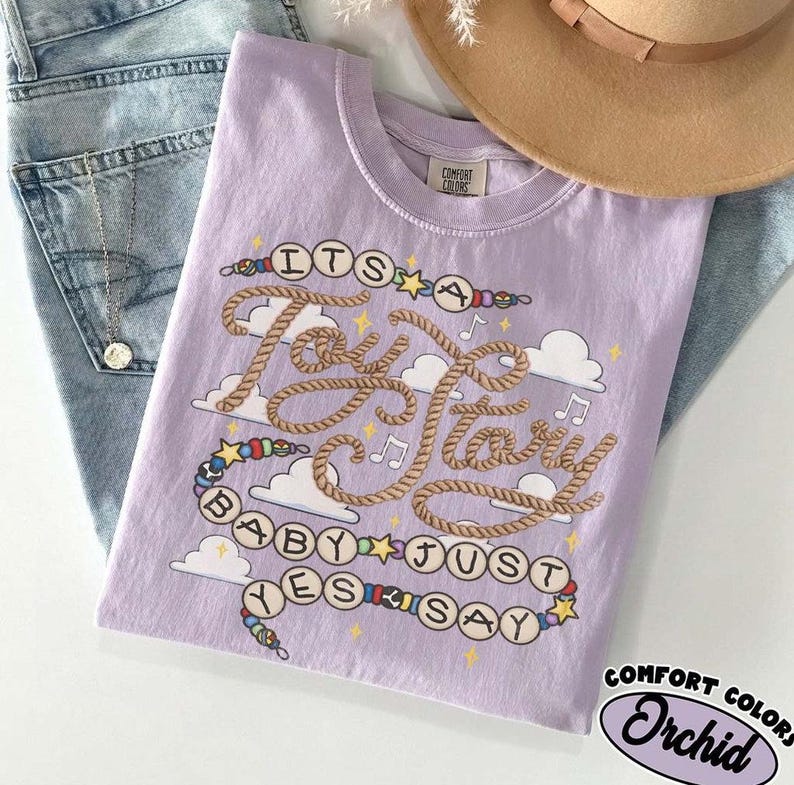 May include: A light purple t-shirt featuring a graphic design with the text "ITS A Toy Story BABY JUST YES SAY" written in rope-style lettering and bead-like circles. The design includes white clouds, small stars, and musical notes.