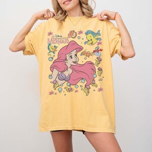 May include: Yellow t-shirt featuring a graphic of Ariel from Disney's The Little Mermaid. The design includes Ariel with pink hair, Flounder, and other sea creatures. The shirt has short sleeves and a crew neck.