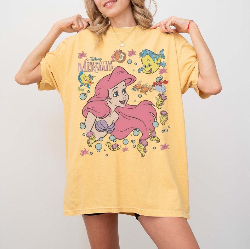 May include: Yellow t-shirt featuring a graphic of Ariel from Disney's The Little Mermaid. The design includes Ariel with pink hair, Flounder, and other sea creatures. The shirt has short sleeves and a crew neck.