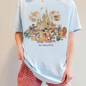 May include: Light blue t-shirt featuring a colorful graphic of Disney characters, including Mickey Mouse, Donald Duck, and Cinderella's castle. The text "Magic Kingdom Walt Disney World" is printed below the graphic.