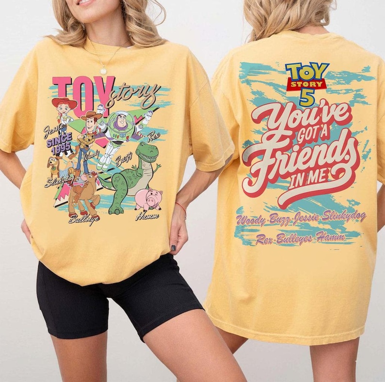 Pode incluir: Uma t-shirt amarela com gr&aacute;ficos das personagens de Toy Story na frente e nas costas. A frente mostra a Jessie, o Woody, o Buzz Lightyear, o Slinky Dog, o Bullseye, o Rex e o Hamm com texto. As costas apresentam o log&oacute;tipo Toy Story 5.