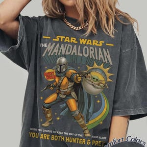 May include: A charcoal gray short-sleeve t-shirt featuring a graphic of The Mandalorian and Grogu. The print includes the text "STAR WARS THE MANDALORIAN" and "WHEN YOU CHOOSE TO WALK THE WAY OF THE MANDALORE YOU ARE BOTH HUNTER & PREY".