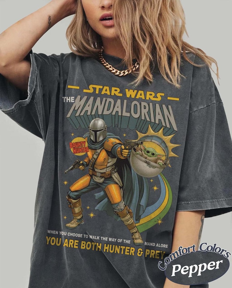 May include: A charcoal gray short-sleeve t-shirt featuring a graphic of The Mandalorian and Grogu. The print includes the text "STAR WARS THE MANDALORIAN" and "WHEN YOU CHOOSE TO WALK THE WAY OF THE MANDALORE YOU ARE BOTH HUNTER & PREY".