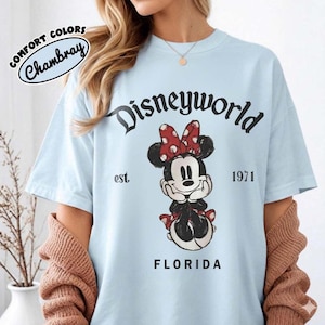 May include: Light blue t-shirt with a Minnie Mouse graphic. The graphic includes the text "Disneyworld" above the character, "est. 1971" and "Florida" below. The shirt is labeled "Comfort Colors Chambray".