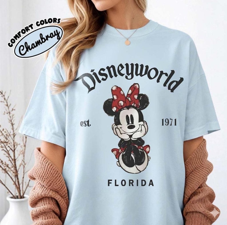 May include: Light blue t-shirt with a Minnie Mouse graphic. The graphic includes the text "Disneyworld" above the character, "est. 1971" and "Florida" below. The shirt is labeled "Comfort Colors Chambray".