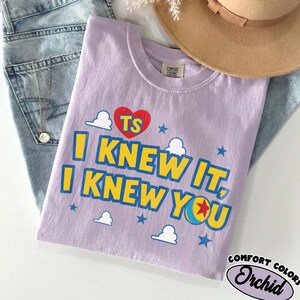 Puede incluir: Una camiseta de color morado claro con un coraz&oacute;n rojo que lleva las letras TS, el texto I KNEW IT, I KNEW YOU en amarillo con bordes azules, y elementos gr&aacute;ficos que incluyen nubes blancas, estrellas azules y una pelota con una estrella roja.