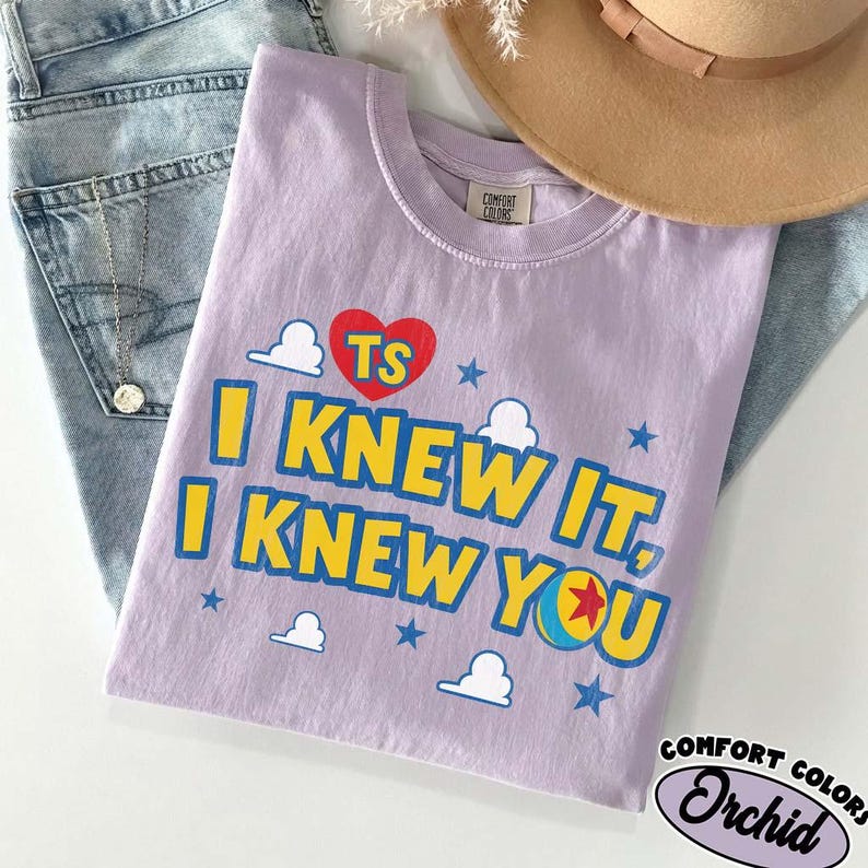 Puede incluir: Una camiseta de color morado claro con un coraz&oacute;n rojo que lleva las letras TS, el texto I KNEW IT, I KNEW YOU en amarillo con bordes azules, y elementos gr&aacute;ficos que incluyen nubes blancas, estrellas azules y una pelota con una estrella roja.