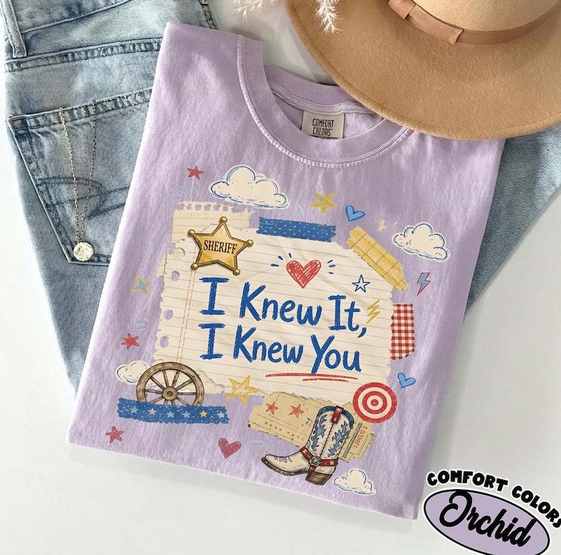 Op de afbeelding: Een lichtpaars t-shirt met korte mouwen en een afbeelding van een gelinieerde notitieblokpagina met de tekst I Knew It, I Knew You. Het ontwerp bevat een gouden sheriffster, een cowboylaars, een wagenwiel, wolken, hartjes en sterren.